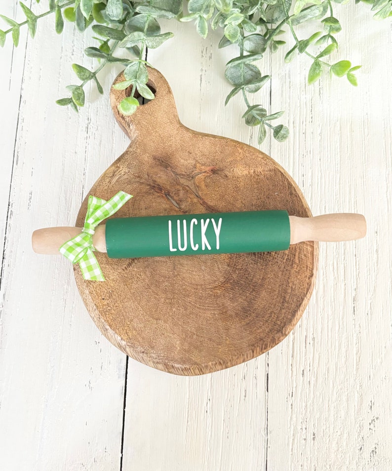 May include: Green rolling pin with a white "LUCKY" inscription on a wooden chopping board with a green and white gingham ribbon.