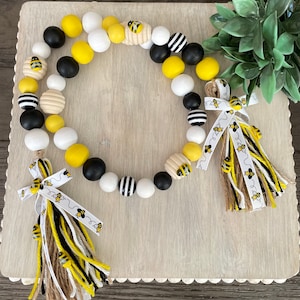 May include: A black, white, and yellow beaded garland with bee accents and two tassels. The garland is made of wooden beads and has a rustic farmhouse style.