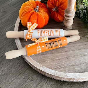 May include: Two wooden rolling pins decorated with orange and white gingham ribbon. One rolling pin is white with black text that says "Pumpkin Pie". The other rolling pin is orange with white polka dots and black text that says "Pumpkin Pie". The rolling pins are on a wooden tray with two orange pumpkins.