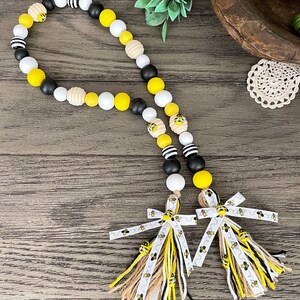May include: A black, white, and yellow beaded garland with bee charms and two tassels with bee-themed ribbon. The tassels are made of burlap, black, white, and yellow yarn.