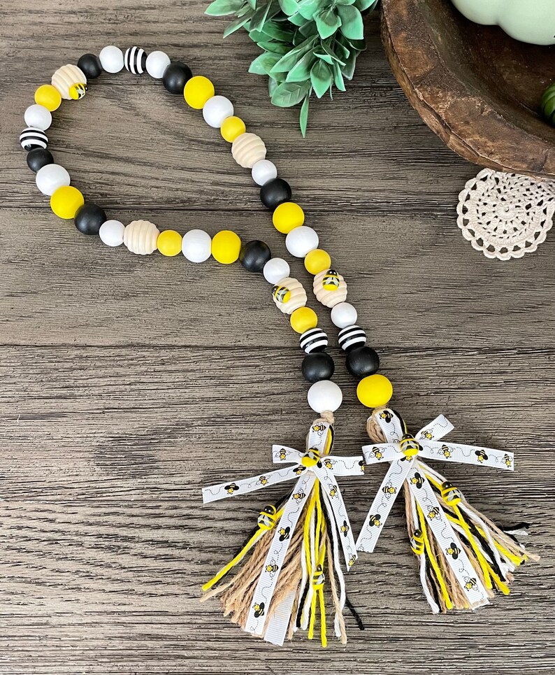 May include: A black, white, and yellow beaded garland with bee charms and two tassels with bee-themed ribbon. The tassels are made of burlap, black, white, and yellow yarn.