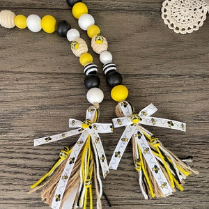 May include: A beaded garland with black, white, and yellow wooden beads, featuring a bee design. The garland has two tassels with white ribbon and brown, black, and white yarn.