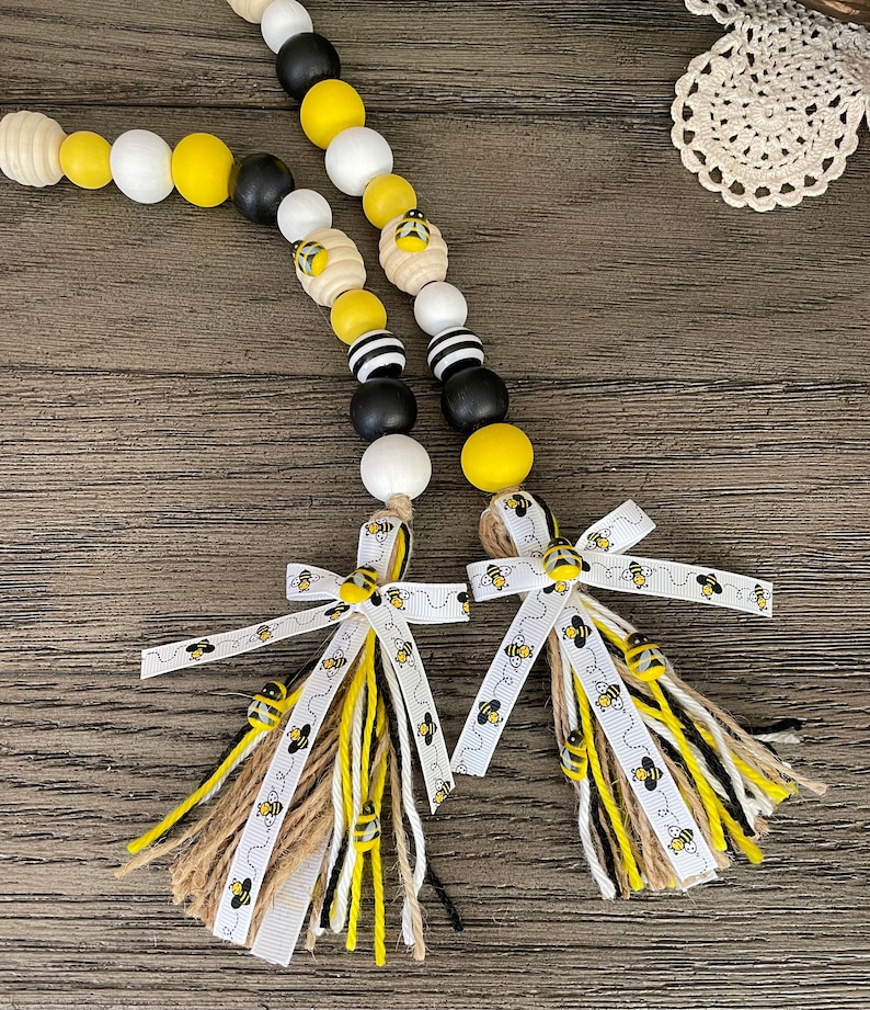 May include: A beaded garland with black, white, and yellow wooden beads, featuring a bee design. The garland has two tassels with white ribbon and brown, black, and white yarn.