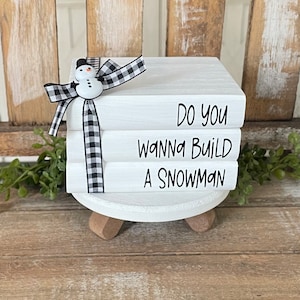 May include: A stack of three white books with black text that reads "Do you wanna build a snowman". The books are tied together with a black and white checkered ribbon and a small snowman figurine is on top.