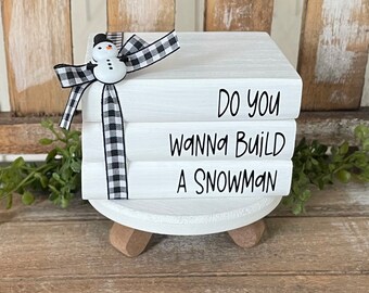 Könnte beinhalten: Ein Stapel von drei weißen Büchern mit schwarzem Text, der "Do you wanna build a snowman" lautet. Die Bücher sind mit einem schwarz-weiß karierten Band zusammengebunden und obenauf befindet sich eine kleine Schneemannfigur.