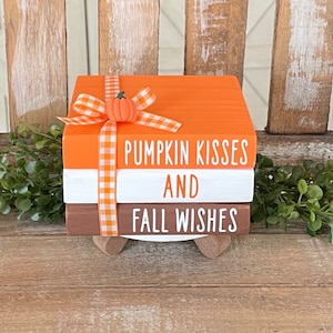 May include: A set of three stacked wooden blocks with a fall theme. The top block is orange and says "Pumpkin Kisses". The middle block is white and says "And". The bottom block is brown and says "Fall Wishes". The blocks are decorated with a white and orange gingham ribbon and a small orange pumpkin.