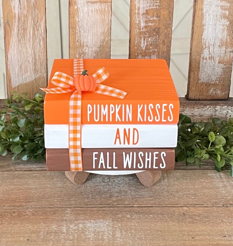 May include: A set of three stacked wooden blocks with a fall theme. The top block is orange and says "Pumpkin Kisses". The middle block is white and says "And". The bottom block is brown and says "Fall Wishes". The blocks are decorated with a white and orange gingham ribbon and a small orange pumpkin.