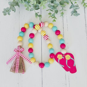 May include: A colorful beaded garland with pink, yellow, blue, and white beads. The garland has a pink and white gingham bow with a brown tassel and a pink wooden flip-flop charm.