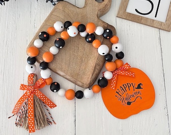 May include: A decorative garland of round wooden beads in black, white, and orange, featuring a jute tassel with an orange polka-dot ribbon and a wooden pumpkin-shaped tag printed with the text Happy Halloween and a witch hat.