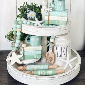 May include: A white tiered tray decorated with a beach theme. The top tier features a stack of books with the words "Beach Please", "Wish Upon A Starfish", and a mug that says "Vitamin Sea". The bottom tier has a jar that says "Surf", a wooden scoop that says "Message in a Bottle", and two rolling pins that say "Ocean Waves" and "Beach Bum".