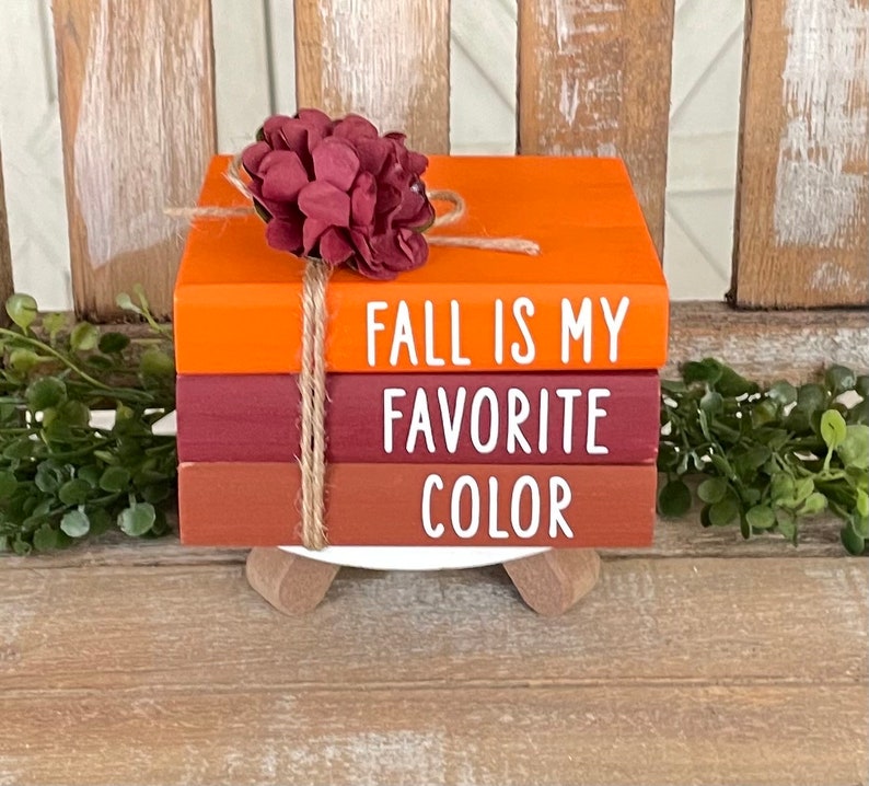 May include: A stack of three faux books with a fall theme. The top book is orange and says "Fall is my favorite color". The middle book is burgundy and says "Favorite". The bottom book is brown and says "Color".
