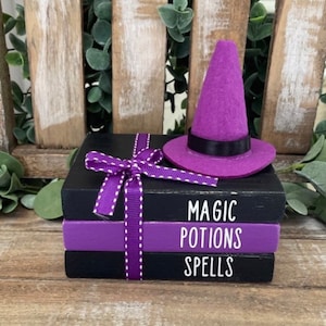 Puede incluir: Una pila de tres bloques de madera negros y morados con las palabras "Magic", "Potions" y "Spells" escritas en blanco. Un sombrero de bruja morado se sienta encima de los bloques.