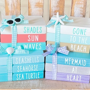 May include: A set of two stacked wooden book-shaped boxes decorated with summer-themed words and images. The boxes are painted in shades of blue, green, white, and pink. The left box has the words "Shades Sun Waves Seashells Seahorse Sea Turtle" and is decorated with a pair of sunglasses and a seashell. The right box has the words "Gone To The Beach Mermaid At Heart" and is decorated with a starfish and a mermaid.