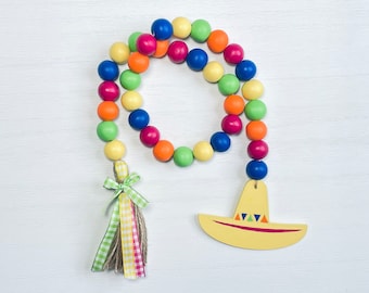 May include: A colorful wooden bead garland with a sombrero pendant. The garland features a variety of painted wooden beads in shades of blue, green, orange, pink, and yellow. A yellow sombrero with colorful triangles hangs from the garland.
