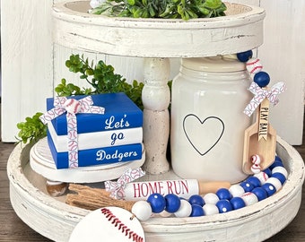 May include: A white tiered tray decorated with a baseball theme. The top tier has a potted plant, a white jar with a heart design, and a wooden baseball bat with the words "Home Run". The bottom tier has a stack of blue and white books with the words "Let's go Dodgers", a wooden baseball, and a white and blue beaded garland.