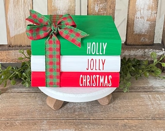 Könnte beinhalten: Ein Stapel aus drei Holzblöcken, die in Rot, Weiß und Grün lackiert sind, mit den Worten "HOLLY", "JOLLY" und "CHRISTMAS", die in Weiß lackiert sind. Die Blöcke sind mit einem rot-grünen karierten Band zusammengebunden.