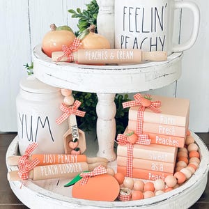 May include: A white tiered tray decorated with peach-themed decor. The top tier features a white mug with black text that reads "Feelin' Peachy", two peach-shaped ceramic ornaments, and a wooden rolling pin with the text "Peaches & Cream". The middle tier features a white jar with the text "Yum", a wooden spoon with the text "Peaches", and a stack of peach-colored books with white text that reads "Farm Fresh Peaches", "Sweet as a Peach". The bottom tier features two wooden rolling pins with the text "Just Peachy Peach Cobbler", a peach-shaped wooden ornament, and a string of peach-colored beads.