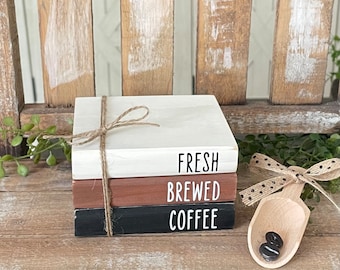 May include: A stack of three rectangular wooden blocks tied with twine, featuring the white text FRESH, BREWED, and COFFEE on white, brown, and black surfaces respectively. A small wooden scoop with a polka dot ribbon holds two dark coffee beans.