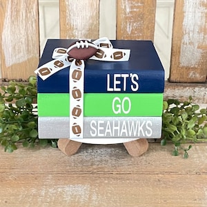 May include: A stack of three wooden blocks decorated with a football theme. The top block is navy blue with the text "LET'S GO" in white. The middle block is green with the text "SEAHAWKS" in white. The bottom block is silver with a white ribbon tied around it. The ribbon has a football pattern.