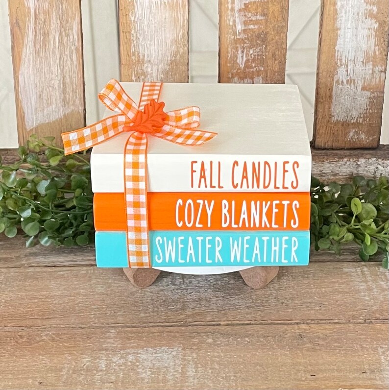 May include: A stack of three white painted wooden blocks with orange and blue accents. The blocks are decorated with the text "Fall Candles", "Cozy Blankets", and "Sweater Weather". The blocks are tied together with an orange and white gingham ribbon and a small orange leaf.