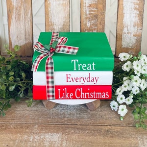 May include: Three stacked books with a red, white, and green color scheme. The top book is green and says "Treat Everyday Like Christmas" in white lettering. The books are decorated with a red and white plaid ribbon with a green bow and holly.