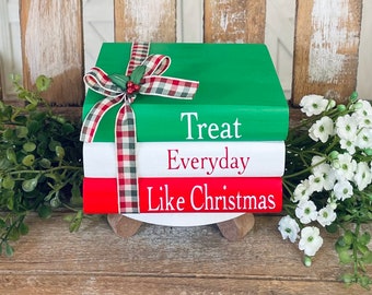 Könnte beinhalten: Drei übereinander gestapelte Bücher in einem rot-weiß-grünen Farbschema. Das oberste Buch ist grün und trägt die Aufschrift "Treat Everyday Like Christmas" in weißen Buchstaben. Die Bücher sind mit einem rot-weiß karierten Band mit einer grünen Schleife und Stechpalme verziert.