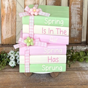 May include: A stack of five wooden blocks painted in green, pink, and white with the text "Spring is in the Air Spring Has Sprung" painted on them. The blocks are decorated with pink and white gingham ribbon and pink and green flowers.