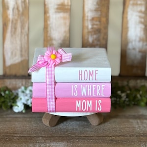 May include: A stack of three pink and white painted books with the words "Home is where Mom is" on the front. The books are tied together with a pink and white gingham ribbon and a pink flower.