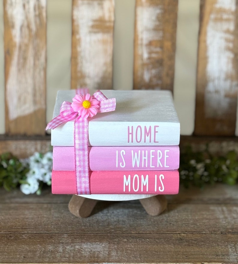May include: A stack of three pink and white painted books with the words "Home is where Mom is" on the front. The books are tied together with a pink and white gingham ribbon and a pink flower.