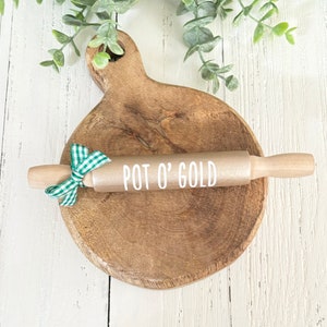 May include: A gold rolling pin with a wooden handle and a white inscription that reads "POT O' GOLD". The rolling pin is on a wooden chopping board with a green and white checked ribbon tied around the handle.
