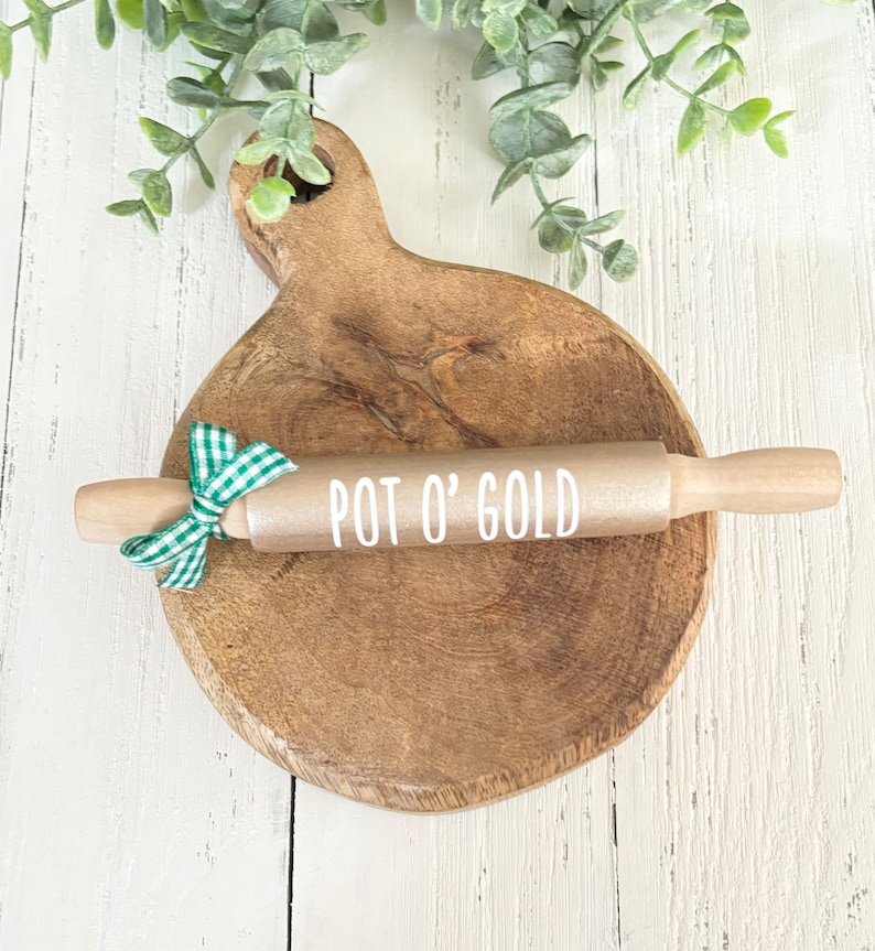 May include: A gold rolling pin with a wooden handle and a white inscription that reads "POT O' GOLD". The rolling pin is on a wooden chopping board with a green and white checked ribbon tied around the handle.