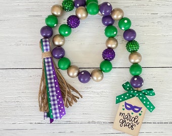 Puede incluir: Una guirnalda de cuentas con temática de Mardi Gras con cuentas verdes, moradas y doradas. La guirnalda tiene un borla con cinta de cuadros verdes y morados y una etiqueta de madera que dice "Mardi Gras" con una máscara morada y corazones.