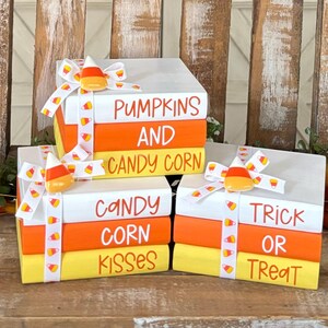 Può includere: Un set di quattro finti libri impilati decorati con nastro e ciondoli di mais dolce arancioni e gialli. I libri sono dipinti di bianco con strisce arancioni e gialle. Il libro in cima dice "Zucca e mais dolce", il libro successivo dice "Baci di mais dolce", il libro successivo dice "Dolcetto o scherzetto".