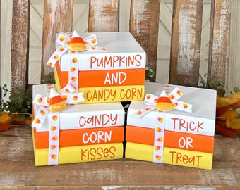 May include: A set of four stacked faux books decorated with orange and yellow candy corn ribbon and candy corn charms. The books are painted white with orange and yellow stripes. The top book reads "Pumpkins and Candy Corn", the next book reads "Candy Corn Kisses", the next book reads "Trick or Treat".
