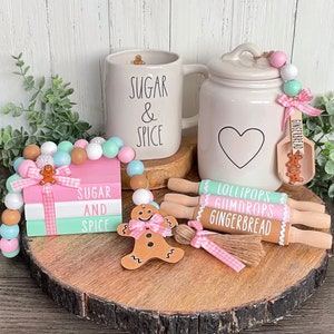 May include: A set of gingerbread themed kitchen decor featuring a white mug with "Sugar & Spice" written on it, a white ceramic jar with a heart design, a stack of wooden blocks with "Sugar and Spice" written on them, a gingerbread man cutout, and three rolling pins with "Lollipops", "Gumdrops", and "Gingerbread" written on them.