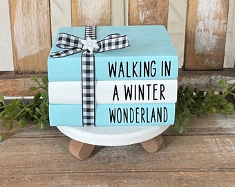 Könnte beinhalten: Ein Stapel aus drei in Blau und Weiß bemalten Büchern mit einem schwarz-weiß karierten Band, das um sie herum gebunden ist. Das oberste Buch trägt die Aufschrift "Walking in a Winter Wonderland".