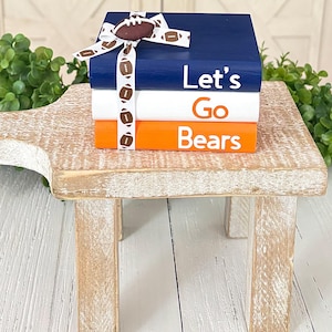 May include: A stack of three decorative books in navy blue, white, and orange, tied with a football-themed ribbon. The books read "Let's Go Bears" in white lettering. The stack sits on a small, rustic wooden stand.