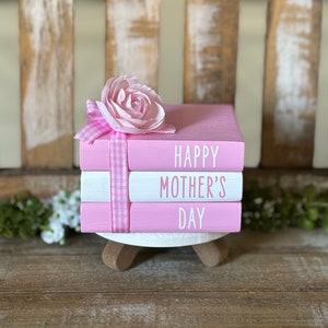 May include: A stack of three pink books with white lettering. The top book says "Happy Mother's Day". A pink flower with a white and pink gingham ribbon is on top of the books.