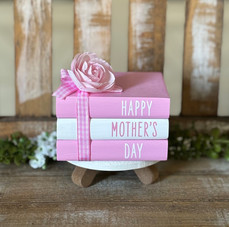 May include: A stack of three pink books with white lettering. The top book says "Happy Mother's Day". A pink flower with a white and pink gingham ribbon is on top of the books.