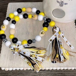 May include: A beaded garland with black, white, yellow, and brown wooden beads. The garland has a bee-themed ribbon with a tassel on each end. The garland is on a wooden surface with a white ceramic bee-themed object in the background.