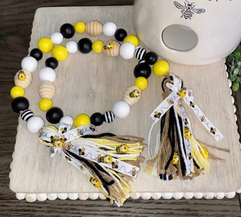 May include: A beaded garland with black, white, yellow, and brown wooden beads. The garland has a bee-themed ribbon with a tassel on each end. The garland is on a wooden surface with a white ceramic bee-themed object in the background.