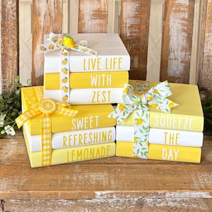 Op de afbeelding: Decoratieve houten boeken in wit en geel, met zinnen als "Live Life With Zest" en "Sweet Refreshing Lemonade". Elke stapel is vastgebonden met een lint met citroenontwerpen. De boeken zijn gerangschikt op een houten oppervlak.