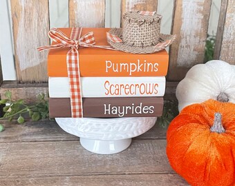 May include: A stack of three wooden blocks with the words "Pumpkins", "Scarecrows", and "Hayrides" painted on them. The blocks are decorated with a burlap and orange ribbon bow and a miniature scarecrow hat.