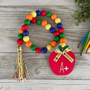 May include: A colorful beaded garland with a red apple charm. The apple has a yellow ribbon with the text "school days" and an A+ grade. The garland has a tassel with the text "school days" on it.