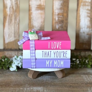 May include: A stack of three pink, white, and purple painted books with the words "I love that you're my mom" on the front. The top book has a white envelope with a small bouquet of flowers on top.