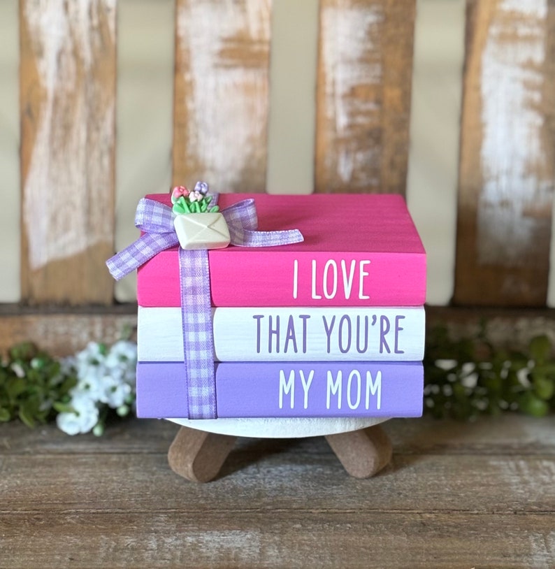 May include: A stack of three pink, white, and purple painted books with the words "I love that you're my mom" on the front. The top book has a white envelope with a small bouquet of flowers on top.