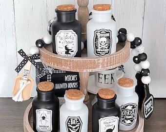 Può includere: Un vassoio di legno a due livelli con un tema di Halloween in bianco e nero. Il vassoio è decorato con perline bianche e nere, un cartello di legno che dice "Witches Haunted Houses" e una tazza che dice "EEK". Ci sono sei bottiglie di vetro con tappi di sughero e etichette che dicono "Witch's Brew", "Poison", "Spider Juice", "The Bat", "Black Cat Hisses" e "Ghost Tears".