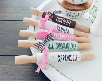 Puede incluir: Seis mini rodillos de amasar con diferentes nombres de sabores y lazos rosas y marrones. Los rodillos de amasar están en una bandeja blanca con una taza de donut blanca y naranja en el fondo. Los sabores son: Chocolate, Vainilla, Fresa, Chip de Chocolate de Menta y Chispas.