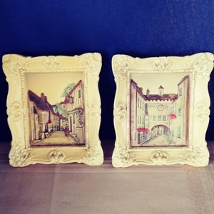 May include: Two framed paintings of European street scenes. The frames are white with ornate details. The paintings are in a vintage style and feature buildings, cobblestone streets, and people.