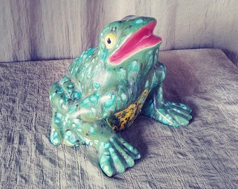 May include: A green ceramic frog figurine with yellow and blue accents. The frog has a wide open mouth and is sitting on a light gray fabric surface.